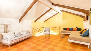 Une pièce de style « loft », avec un plafond haut, des poutres apparentes, un sol à carreaux orange, un canapé, un fauteuil et un lit agrémenté de coussins colorés.