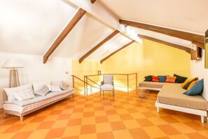 Une pièce de style « loft », avec un plafond haut, des poutres apparentes, un sol à carreaux orange, un canapé, un fauteuil et un lit agrémenté de coussins colorés.
