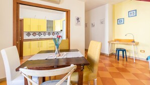 Un coin repas avec une table en bois, des chaises blanches et jaunes, et une cuisine visible à travers une porte ouverte.