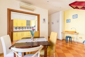 Un coin repas avec une table en bois, des chaises blanches et jaunes, et une cuisine visible à travers une porte ouverte.