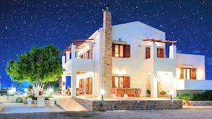 A modern two-story house with a stone chimney, wooden shutters, and a well-lit outdoor area with a bench and potted plants. - Cretan Dream Villa (Apokoronas)