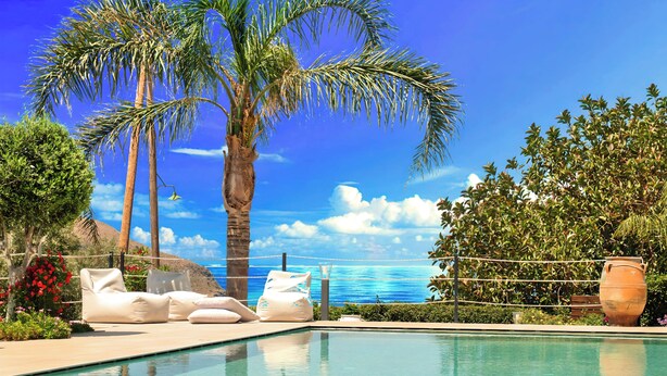 A poolside area with lounge chairs, a palm tree, and a view of the ocean. - Cretan Dream Villa (Apokoronas)