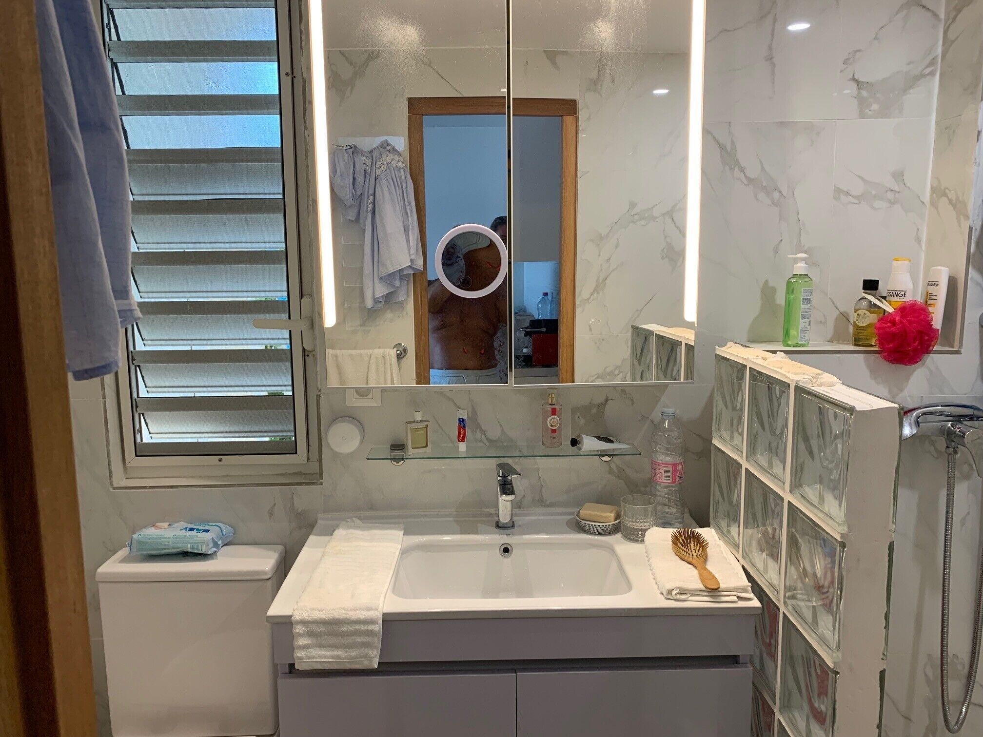 Une salle de bain avec un lavabo blanc, un miroir et une douche. On trouve des articles de toilette sur le lavabo et une serviette est accrochée au mur.