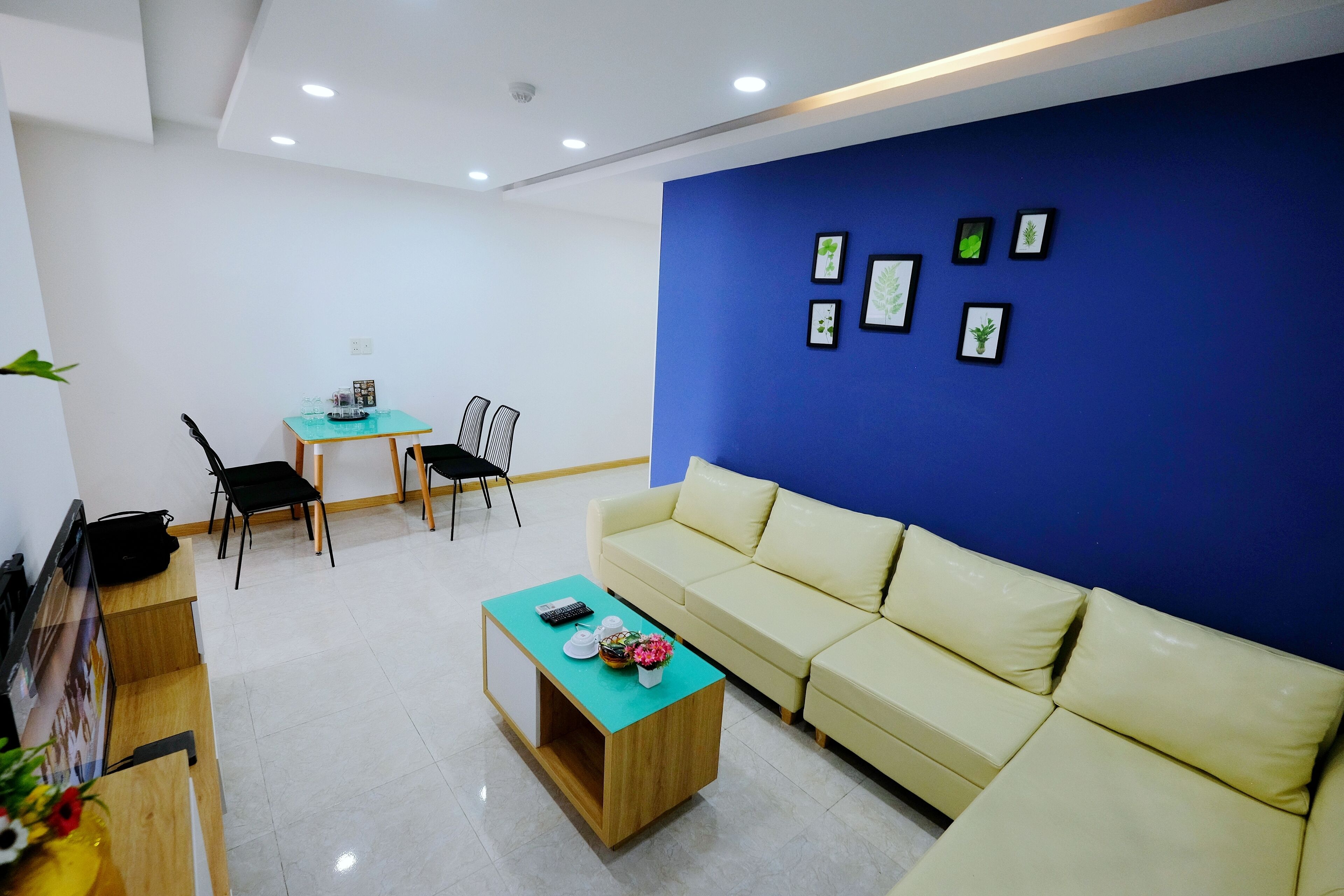 Una sala de estar moderna con un sofá color beige, una mesa de centro y una pared de acento azul. Hay cuadros enmarcados en la pared y una pequeña mesa con sillas.