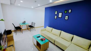 Una sala de estar moderna con un sofá color beige, una mesa de centro y una pared de acento azul. Hay cuadros enmarcados en la pared y una pequeña mesa con sillas.