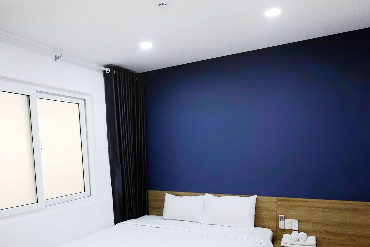 A modern hotel room with a large bed, a wooden headboard, a window with curtains, and a nightstand with a lamp.