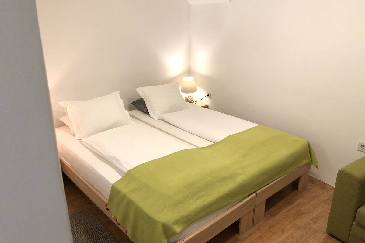 A single bed with a green blanket, white pillows, and a bedside lamp.