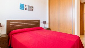 A bedroom with a wooden bed, a red bedspread, a nightstand with a lamp, a wooden wardrobe, and a wall-mounted artwork. - DON PEDRO APARTMENT IN ALCOCEBRE (Alcocéber)