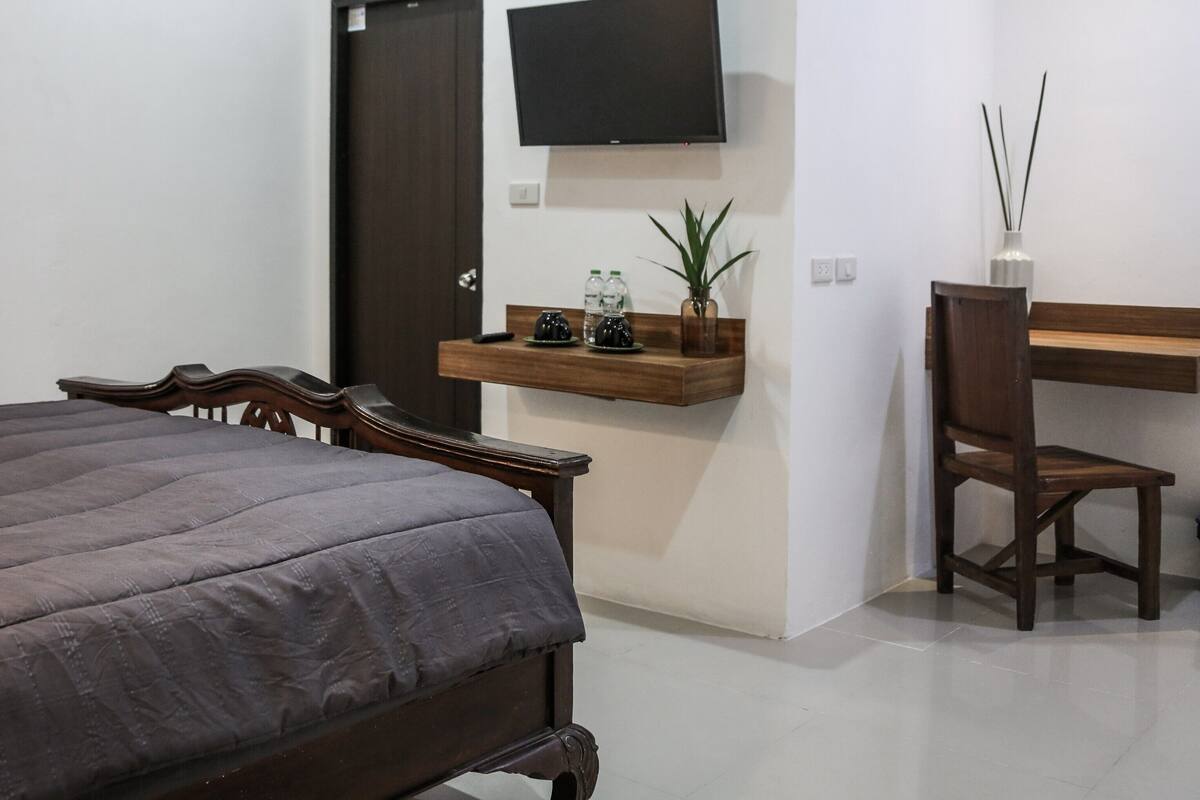 A bedroom with a bed, a wooden nightstand, a desk with a chair, a TV mounted on the wall, and a shelf with decorative items.
