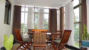 Ein Zimmer mit Holztisch und Stühlen, großen Fenstern mit Vorhängen sowie einem Blumentopf.