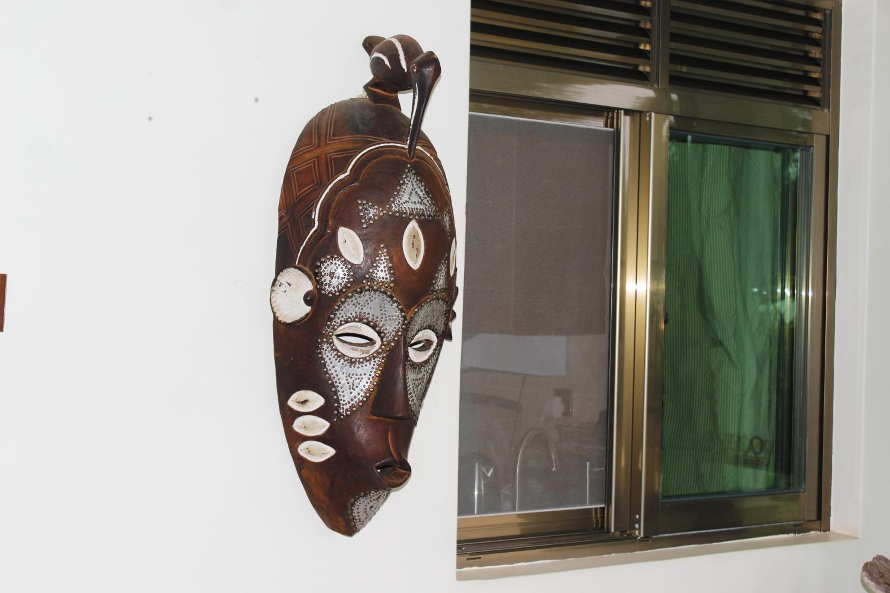 A wall-mounted wooden mask with intricate patterns and a window with a green curtain.