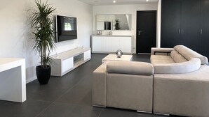 Een moderne woonkamer met een bank, een televisie, een plant en een keuken op de achtergrond.