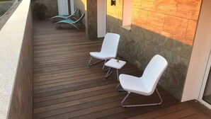 Een houten terras met witte wickerstoelen en een kleine tafel, een stenen muur en een deur op de achtergrond.