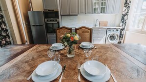 Une table à manger dressée pour quatre personnes, avec de l’argenterie, des assiettes blanches et des verres à vin, un vase rempli de fleurs et une vue sur une cuisine moderne aux armoires grises et équipée d’un lave-linge.