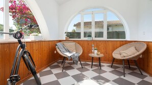 A sunlit balcony with a checkered floor, two wicker chairs, a small round table, and a bicycle.