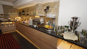 A modern breakfast bar with a stone wall backdrop, a decorative rooster, and a variety of breakfast items including coffee machines and condiments. - Hotel Anika (Neuenburg am Rhein)