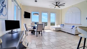 Amplia sala de estar con ventilador de techo, televisor de pantalla plana, mesa de comedor con sillas, sofá blanco y ventanas amplias con vistas al exterior.