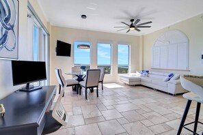A spacious living area with a ceiling fan, a flat-screen TV, a dining table with chairs, a white sofa, and large windows offering a view of the outdoors. - PENTHOUSE SUITE | EXTENDED CEILINGS | ESTERO BAY & GULF VIEWS | SOAKING TUB | LOVERS KEY RESORT (Fort Myers Beach)