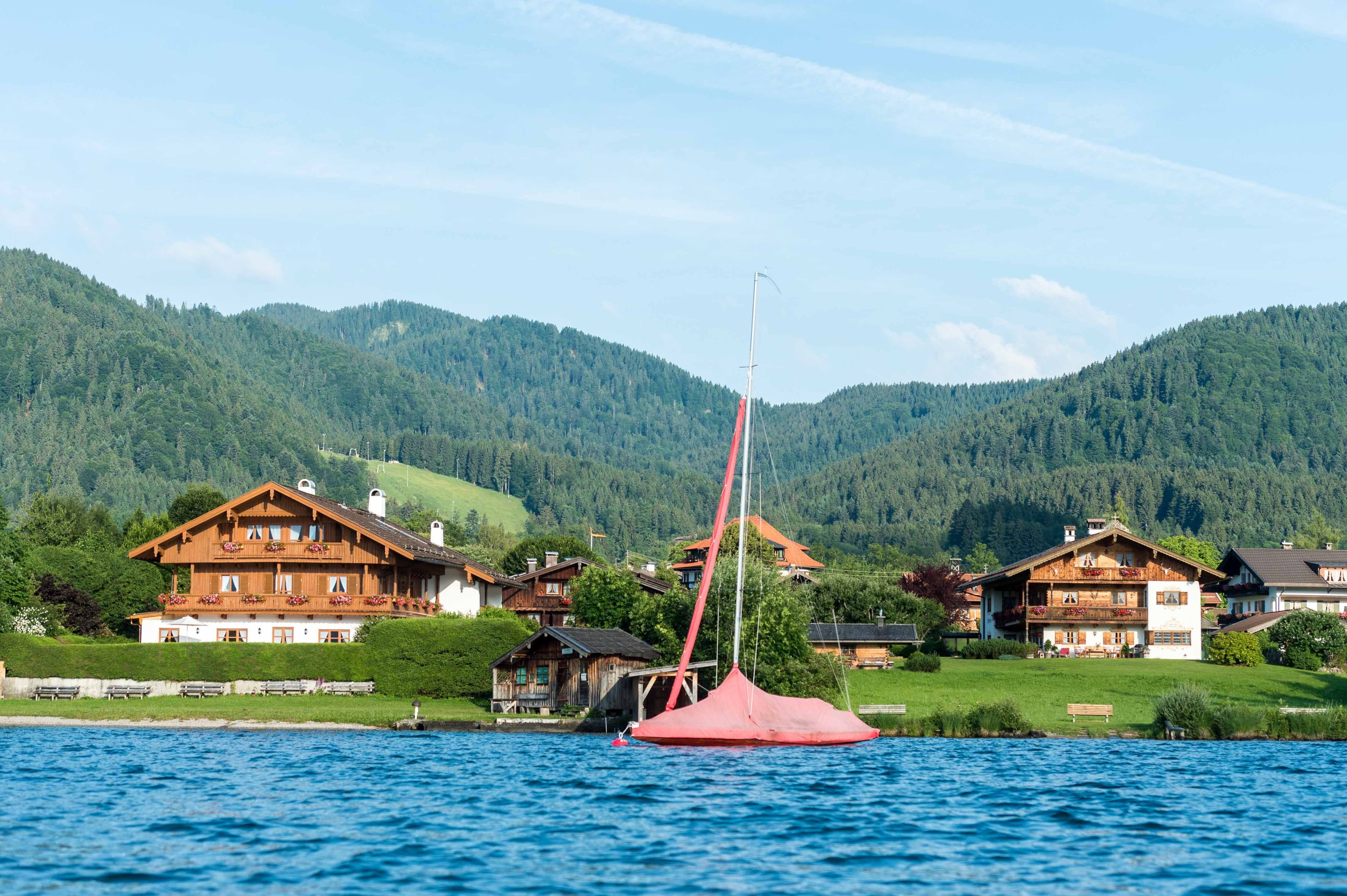 A lakeside property with wooden houses, a red sailboat, and a forested mountain backdrop.