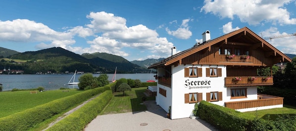 A traditional chalet-style building with a sign reading 'Seerose Ferienwohnungen' by a lake with boats and mountains in the background.