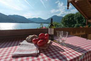 A table with a checkered tablecloth, a bottle of wine, glasses, bread, and tomatoes on a wooden deck overlooking a lake with mountains in the background. - Vacation apartment 2 - Seerose am See - Dream vacation directly on Lake Tegernsee (Bad Wiessee)