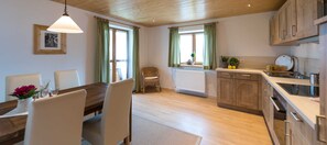 Une cuisine avec des meubles de rangement en bois, une table à manger avec des chaises, un luminaire suspendu et une fenêtre avec des rideaux.