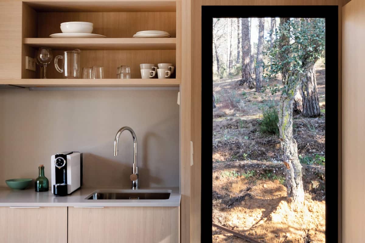 A modern kitchen with wooden cabinets, a sink, and a view of a forest through a glass door.
