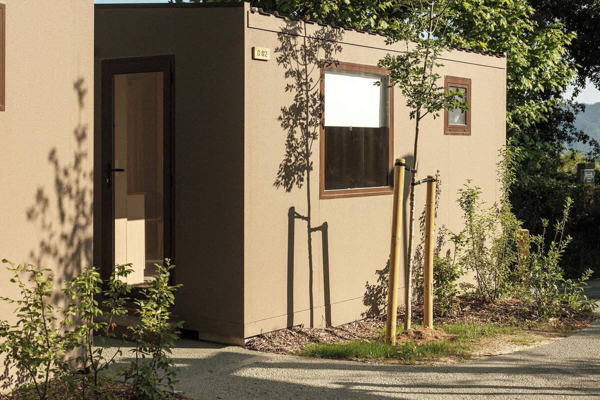 A small, single-story building with a door and a window, surrounded by greenery and a clear sky.