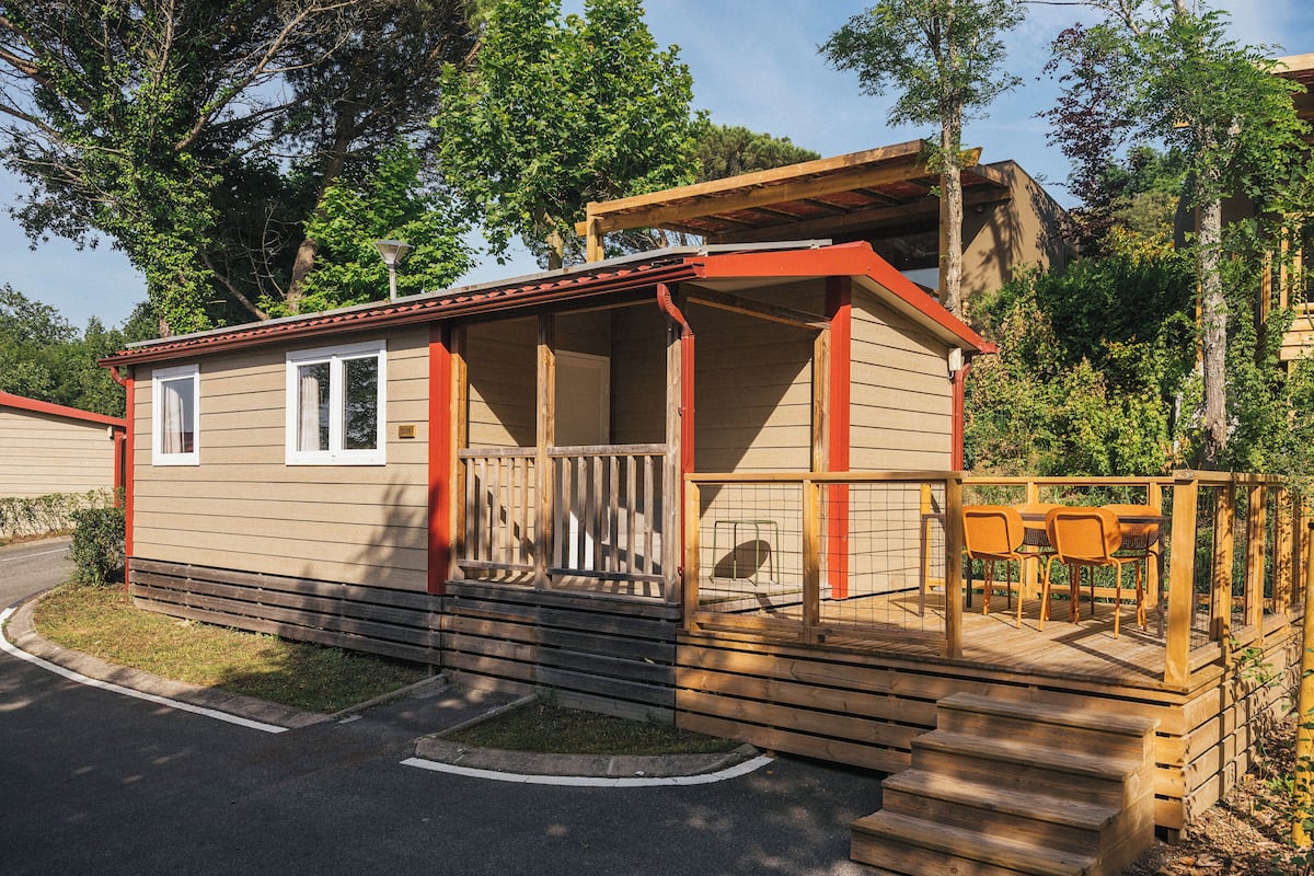 A small, single-story cabin with a porch and a small deck, surrounded by trees.