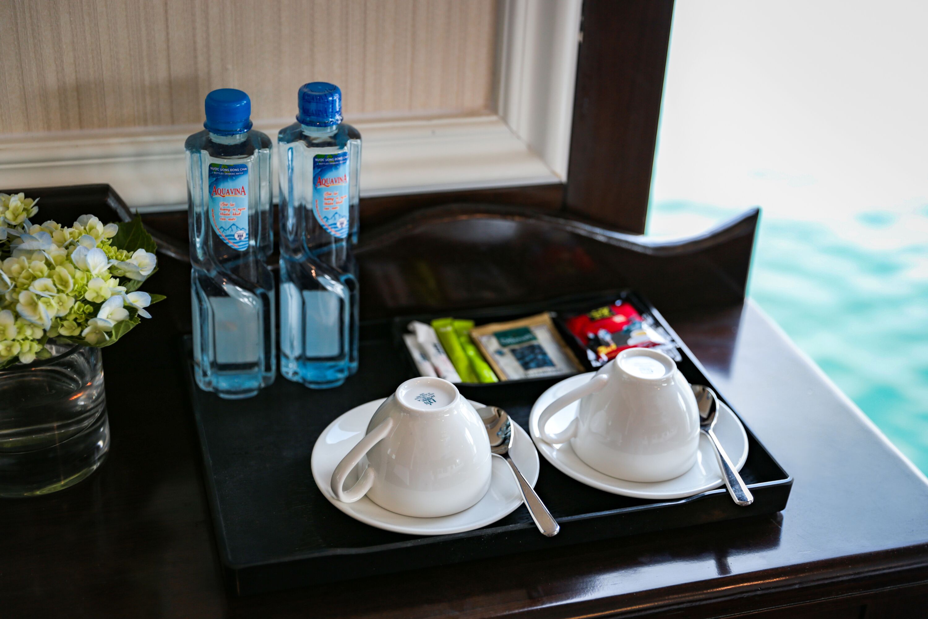Una bandeja con dos tazas blancas de té, una cuchara, dos botellas de agua Aquavina, un jarrón con flores blancas y una bandeja de bocadillos.