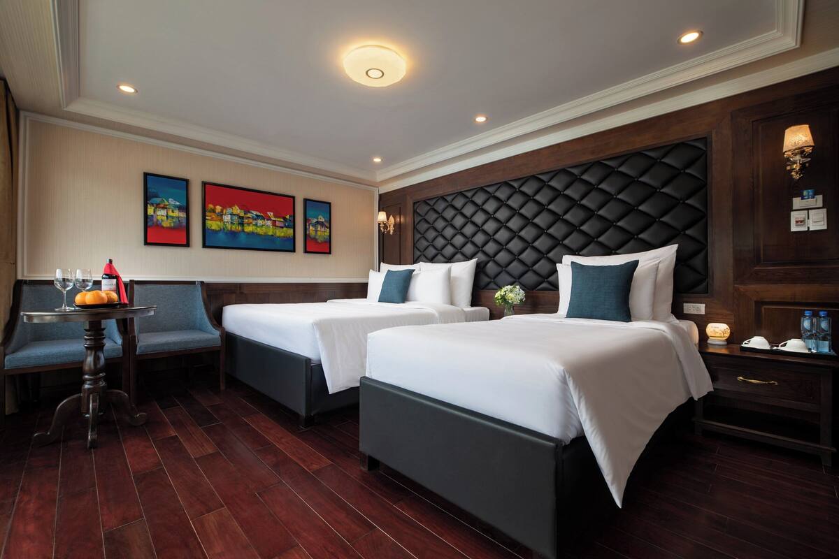 A hotel room with two beds, a small table with glasses, and artwork on the wall.