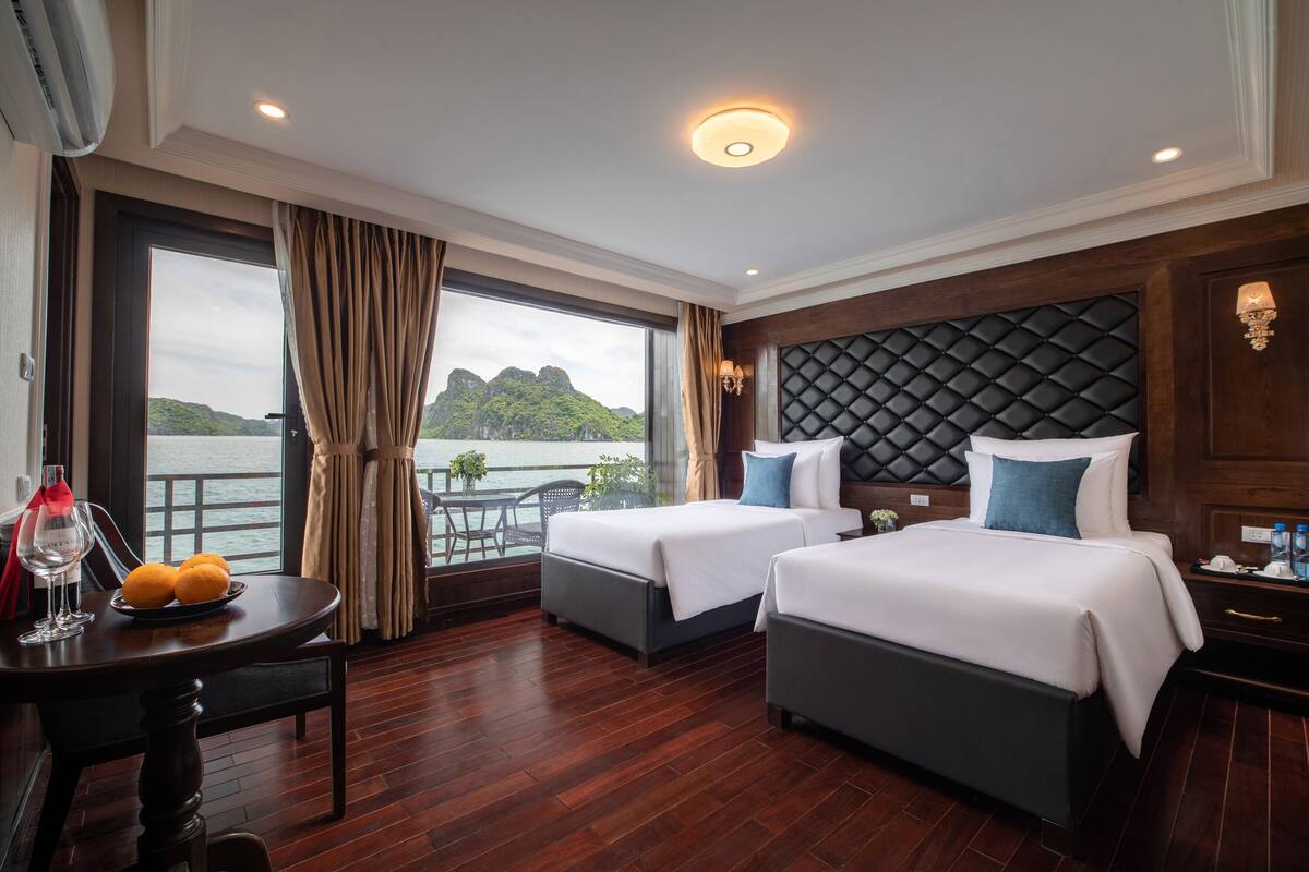 A hotel room with two beds, a dining table with fruits, a view of the sea, and a balcony.