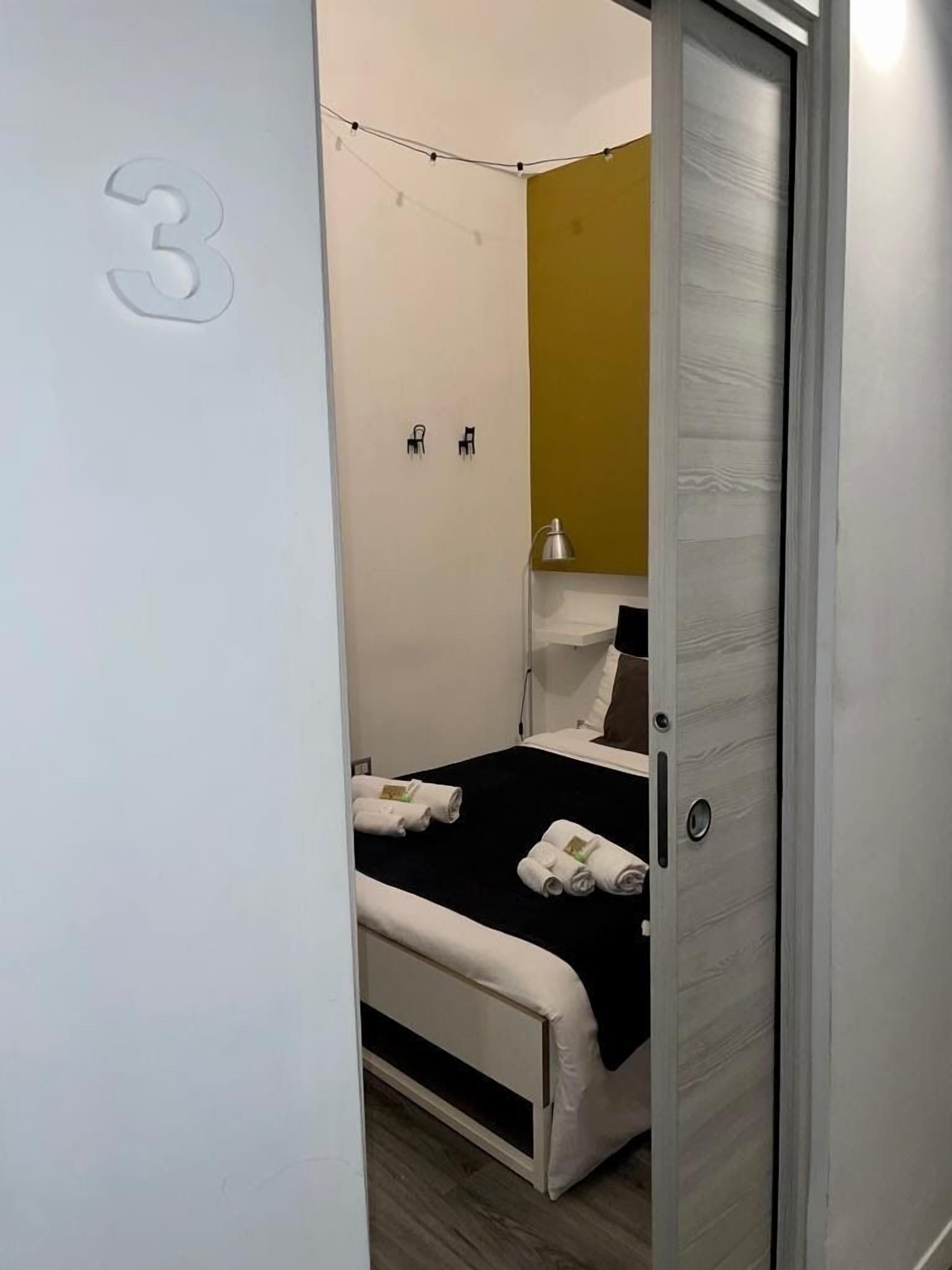 Et hotelværelse med en seng, et natbord, en lampe og en dør med et nummer.
