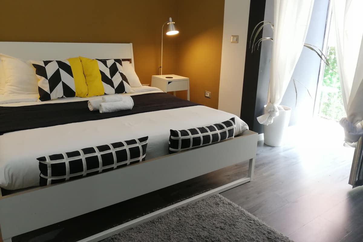 A modern bedroom with a large bed, white and black bedding, a bedside lamp, and a view of greenery outside.