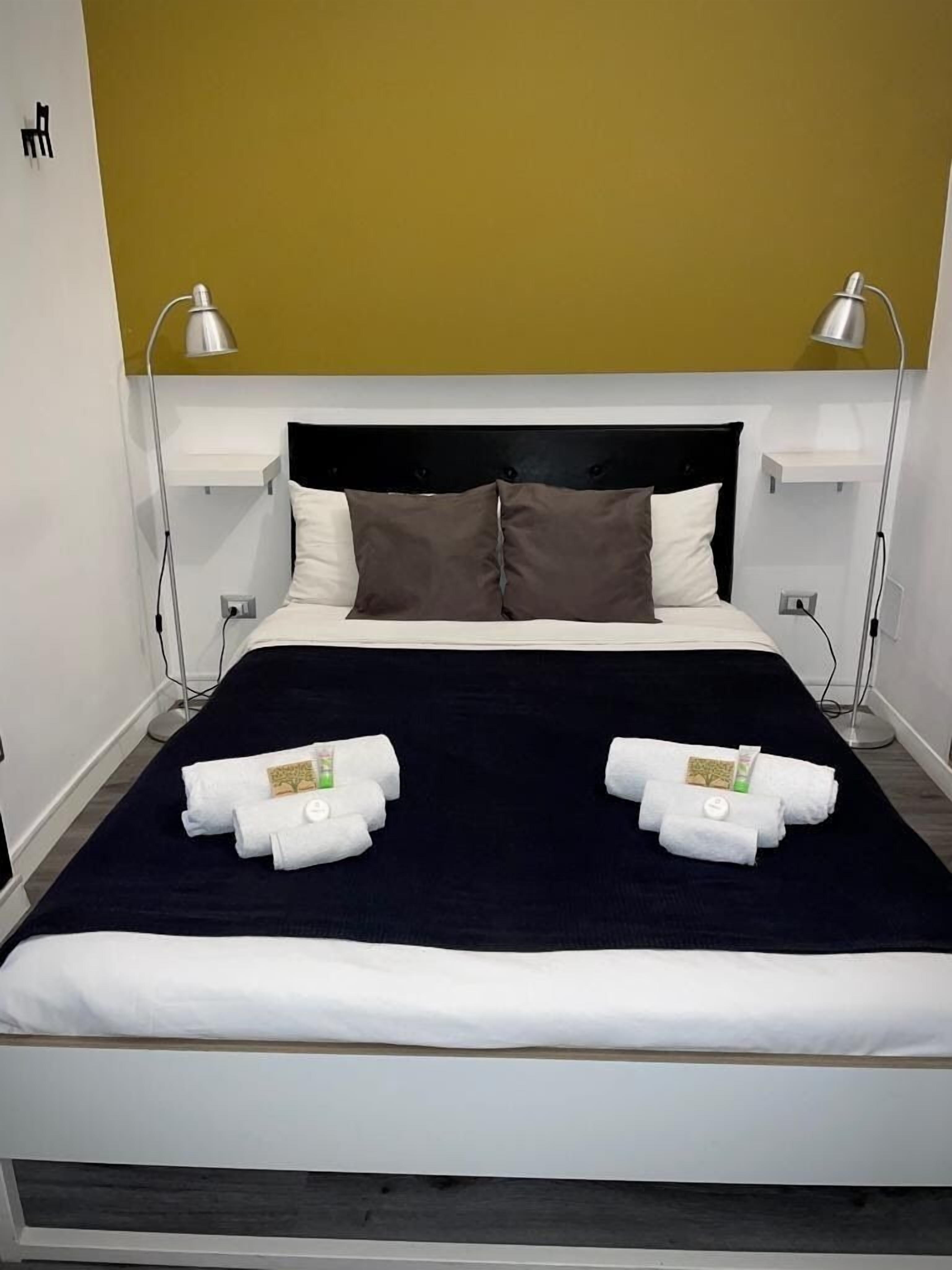 A hotel room with a bed, two bedside lamps, a black headboard, and two towels with toiletries on the bed.