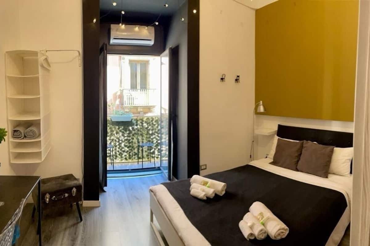 A modern hotel room with a large bed, a small desk, a chair, and a balcony with a view.