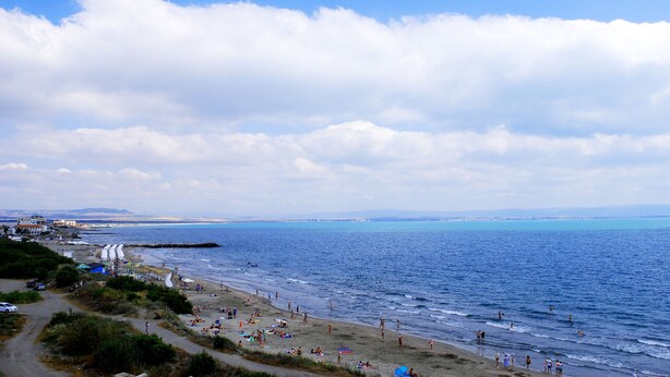 Escena de playa costera con cielo despejado, orilla arenosa y algunos edificios al fondo.