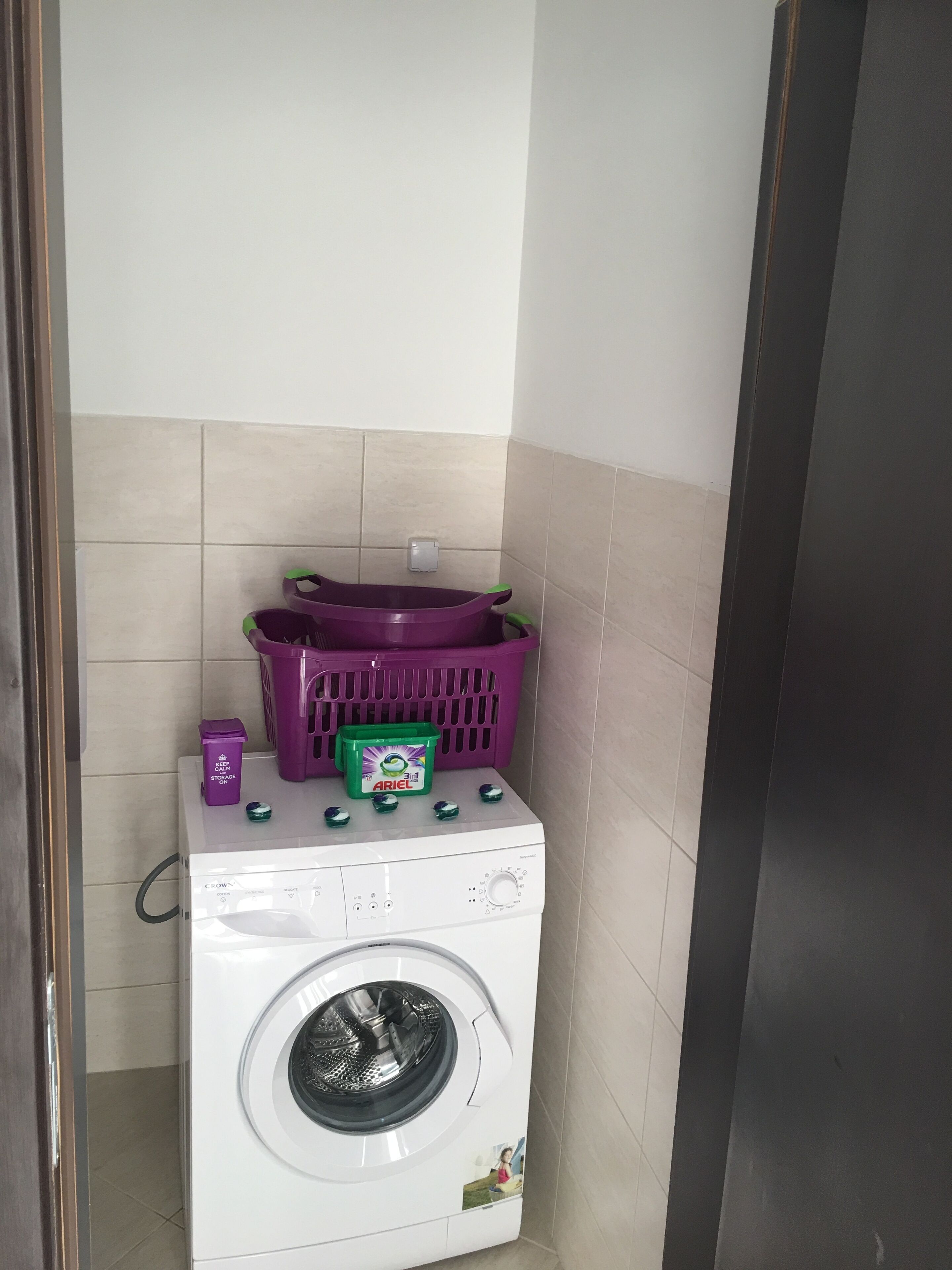 A compact laundry area with a washing machine, a purple basket, and a green detergent box.
