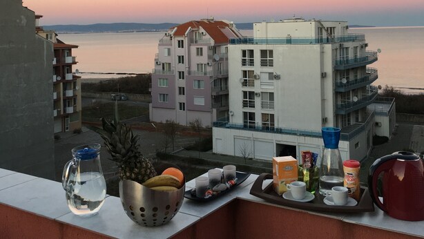 A balcony with a view of the sea and buildings, featuring a tray with cups, a pineapple, and a broken glass pitcher. - Sea Breeze Pomorie (Pomorie)