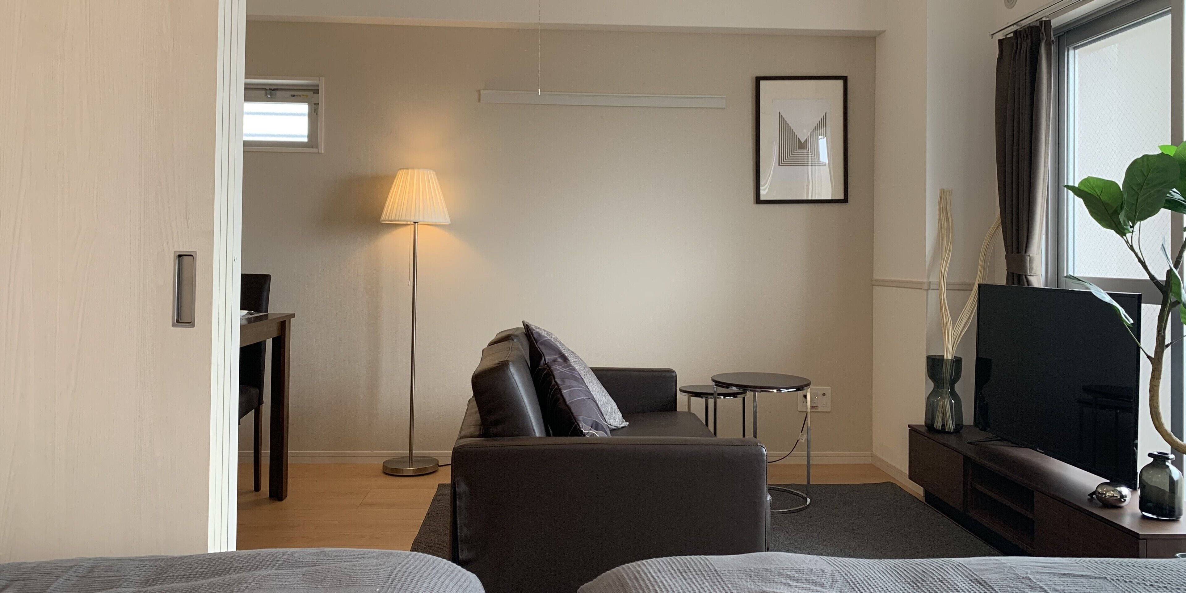 A modern hotel room with a sofa, a small side table, a floor lamp, a TV, and a large window.