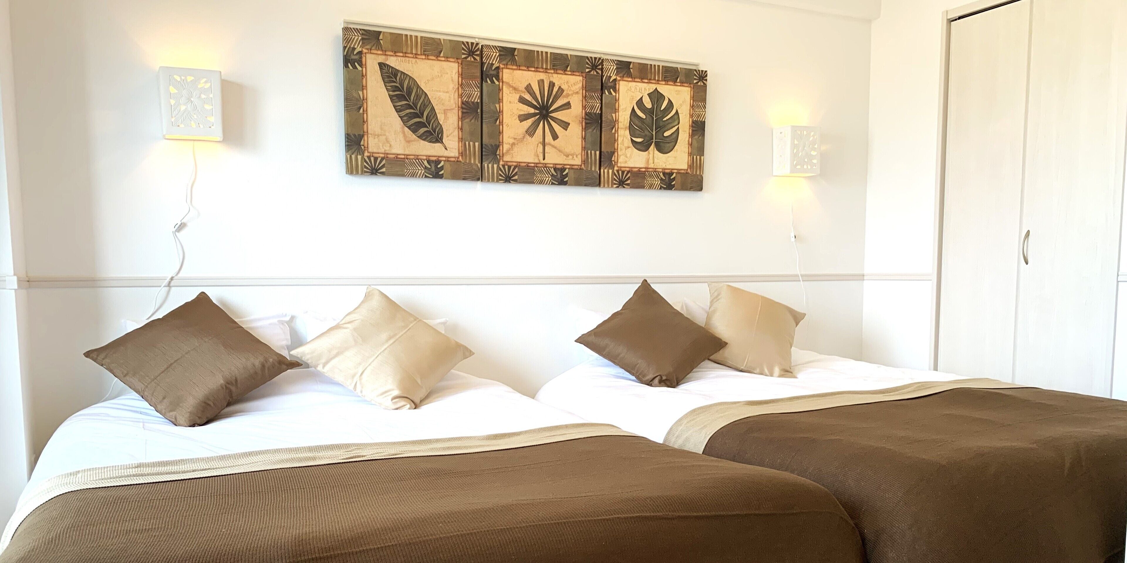 Quarto de hotel com duas camas, roupa de cama bege e castanha, um quadro na parede com motivos de folhas e luminárias de cabeceira.