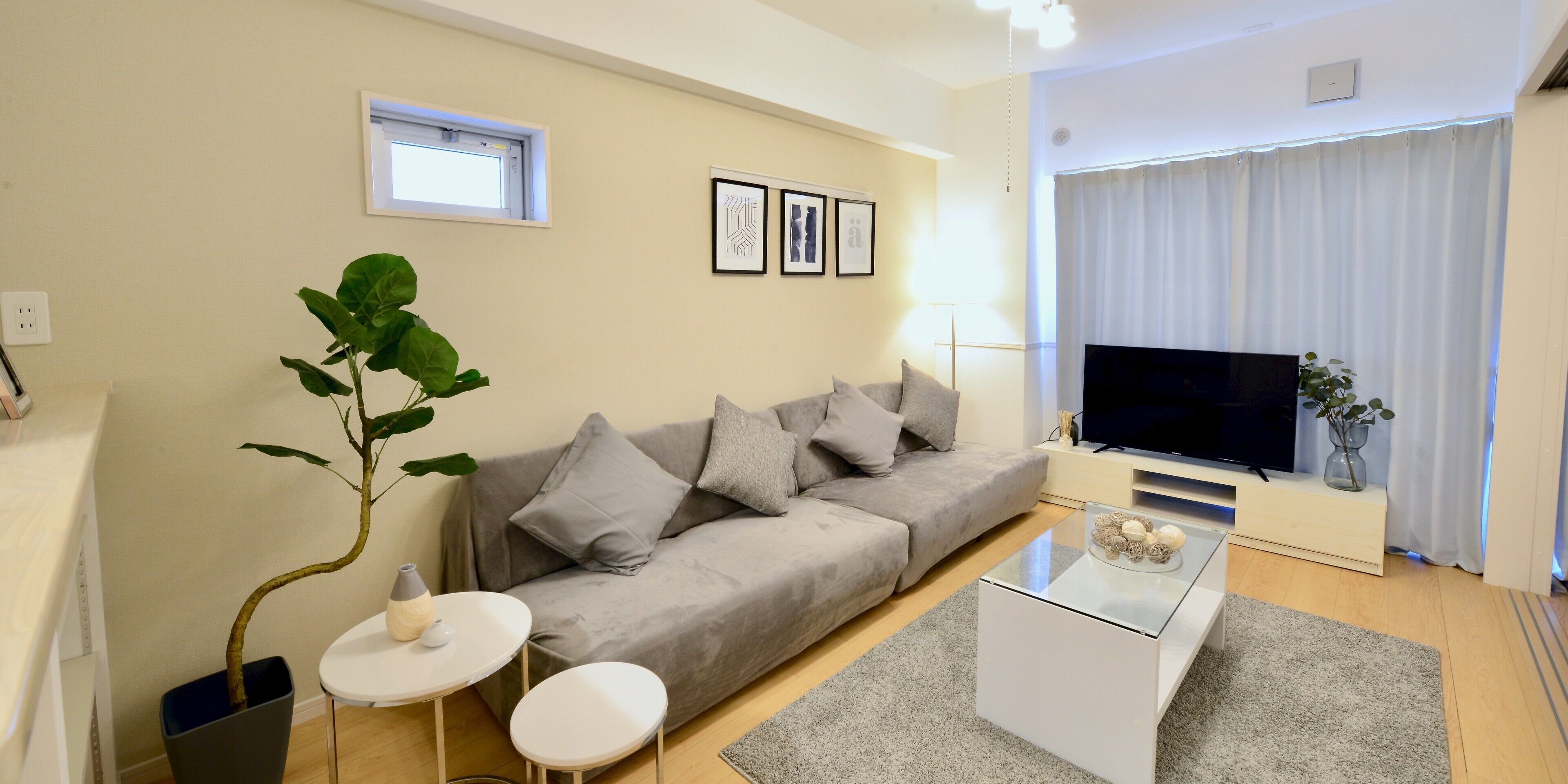 En moderne stue med en grå sofa, et glasbord, et lille hvidt bord og et fjernsyn på et stativ.
