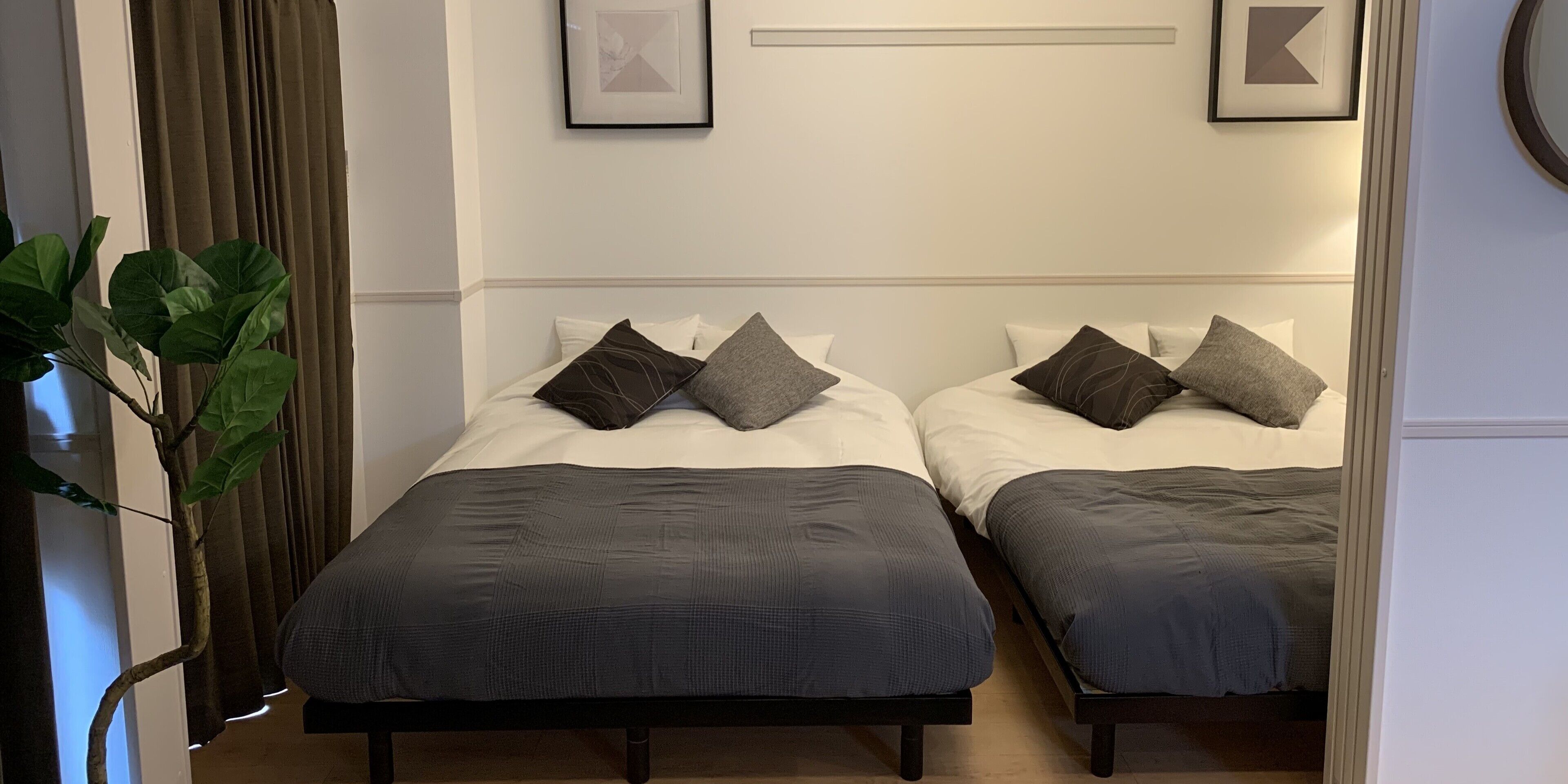 Duas camas de solteiro com roupa de cama escura, um vaso de planta e quadros na parede.