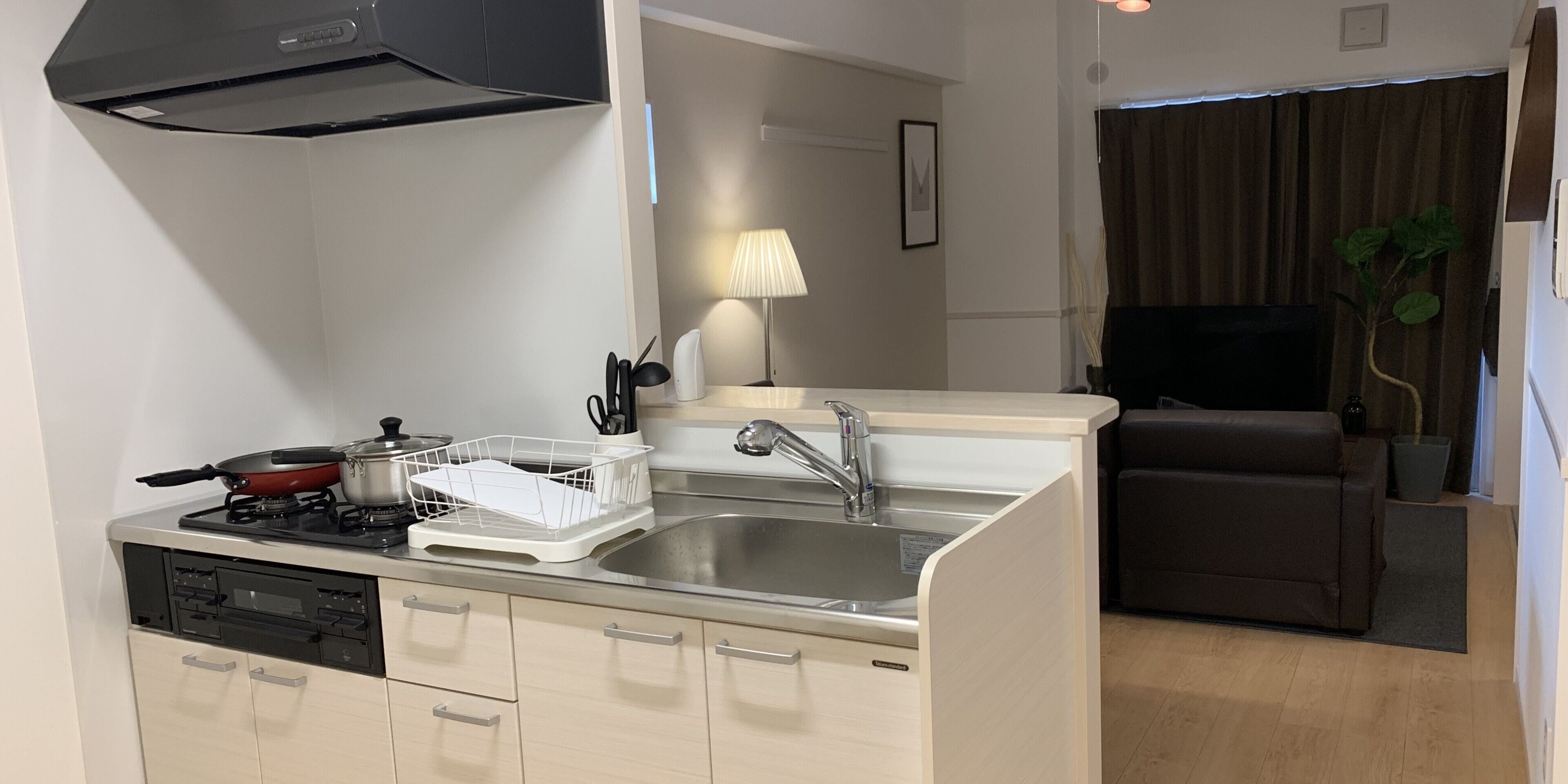A compact kitchen with a stove, sink, and integrated dishwasher. A small dining area with a sofa and a lamp is visible in the background.