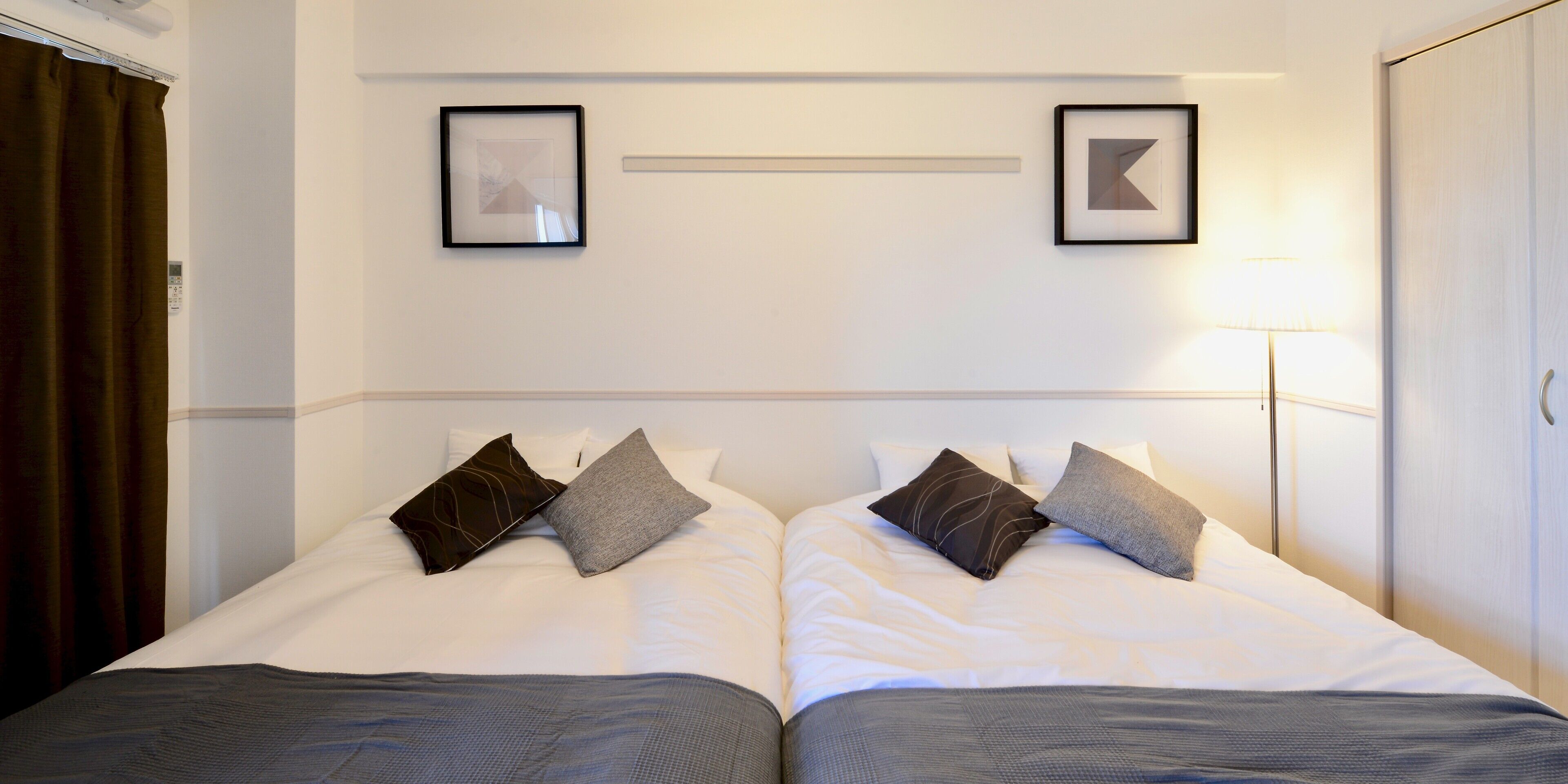 Een hotelkamer met twee bedden, elk voorzien van een grijze plaid en meerdere kussens. Er hangen fotolijstjes aan de muur boven de bedden, een bedlamp en een garderobe.