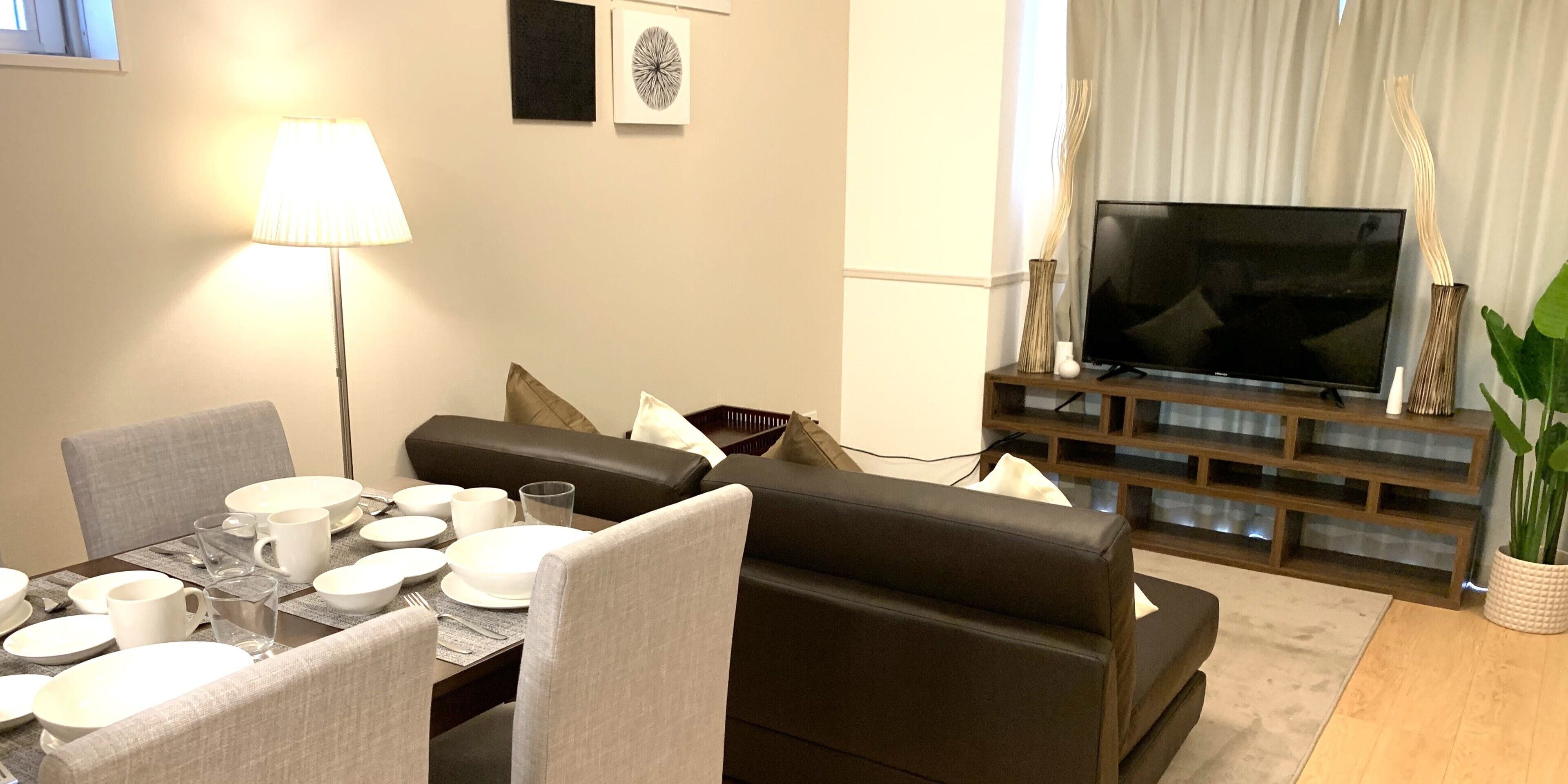 A modern living room with a dining area set for four, a flat-screen TV, and a large potted plant.