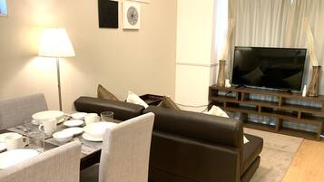 A modern living room with a dining area set for four, a flat-screen TV, and a large potted plant.