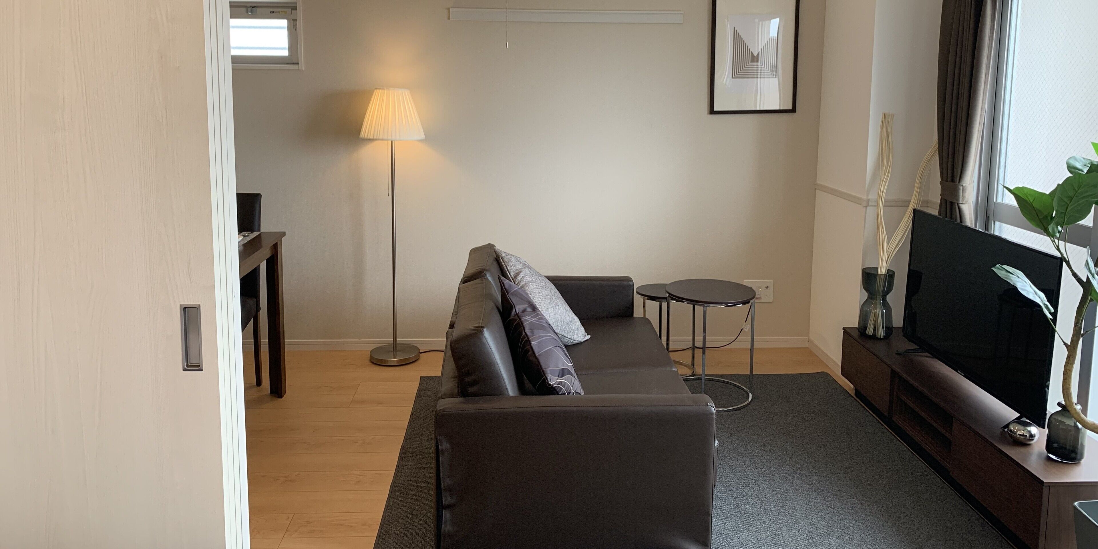 Een moderne woonkamer met een donker leren bank, een staande vloerlamp, een klein rond tafeltje en een flatscreen televisie op een houten standaard.