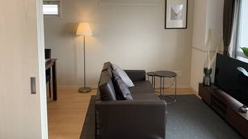 Een moderne woonkamer met een donker leren bank, een staande vloerlamp, een klein rond tafeltje en een flatscreen televisie op een houten standaard.