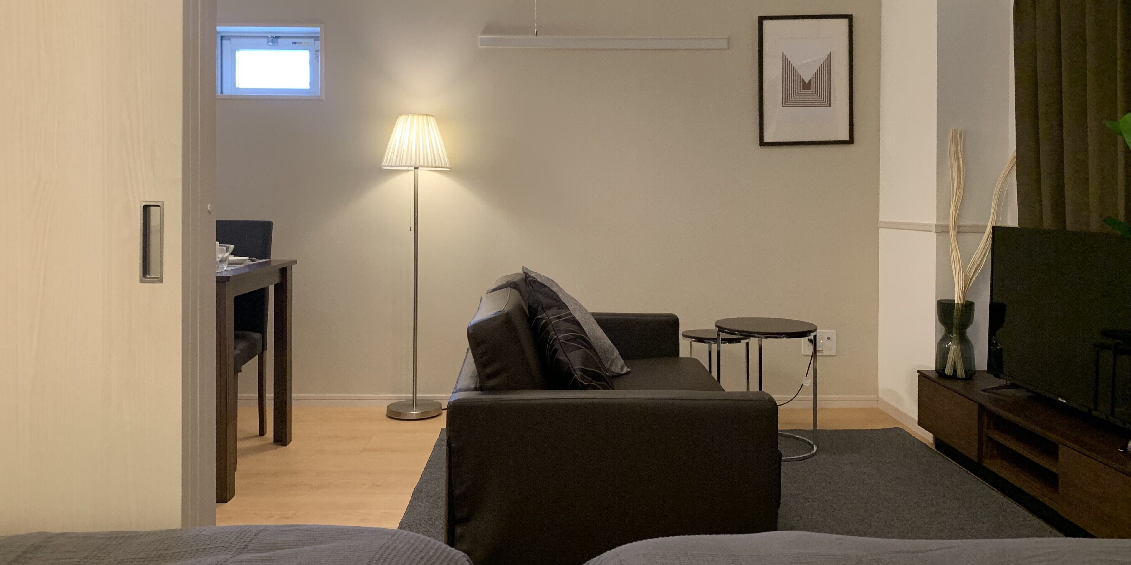 A modern hotel room with a sofa, a small table, a floor lamp, a TV, and a framed picture on the wall.